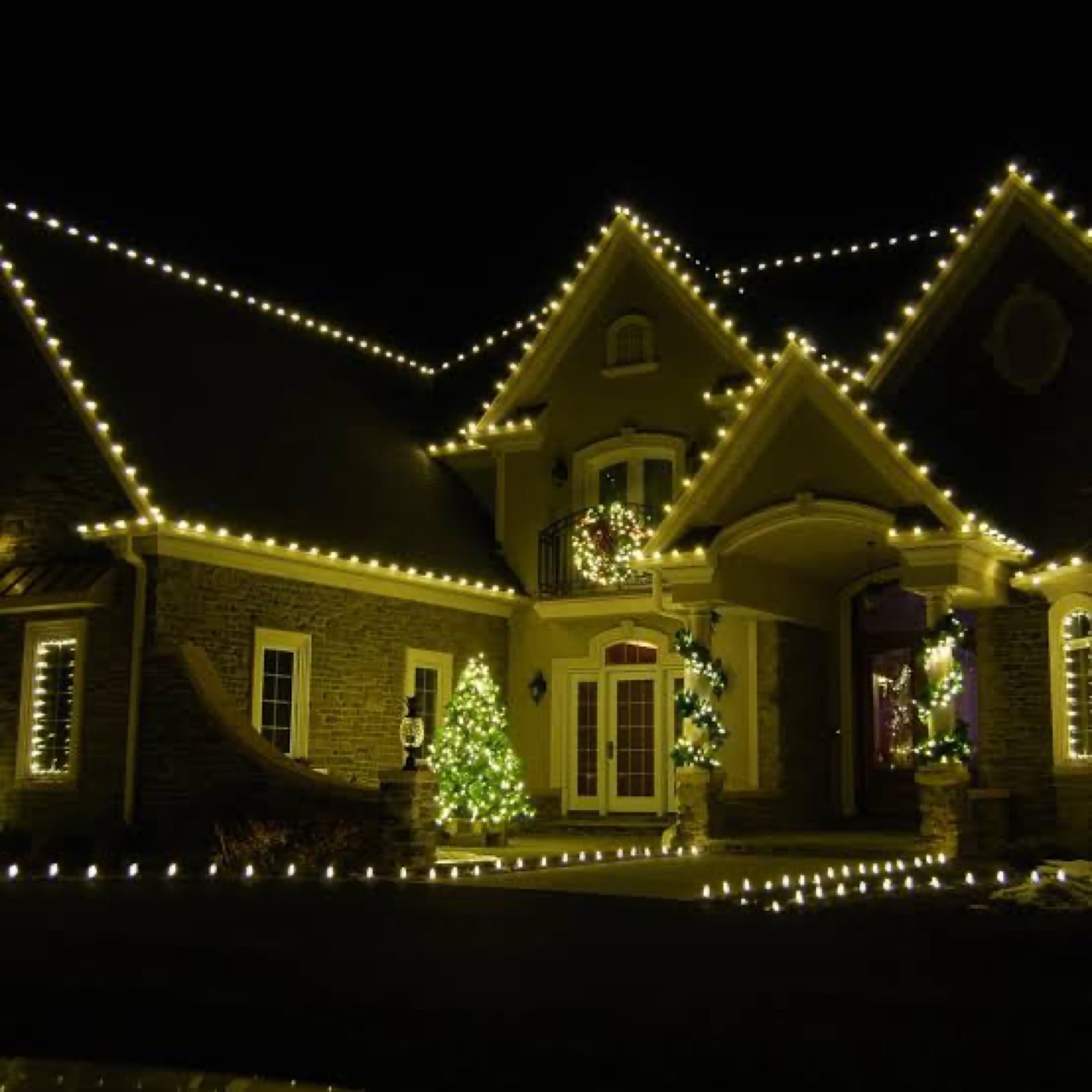 Warm white roofline lights, lit wreaths and illuminated evergreens on a stone house at night