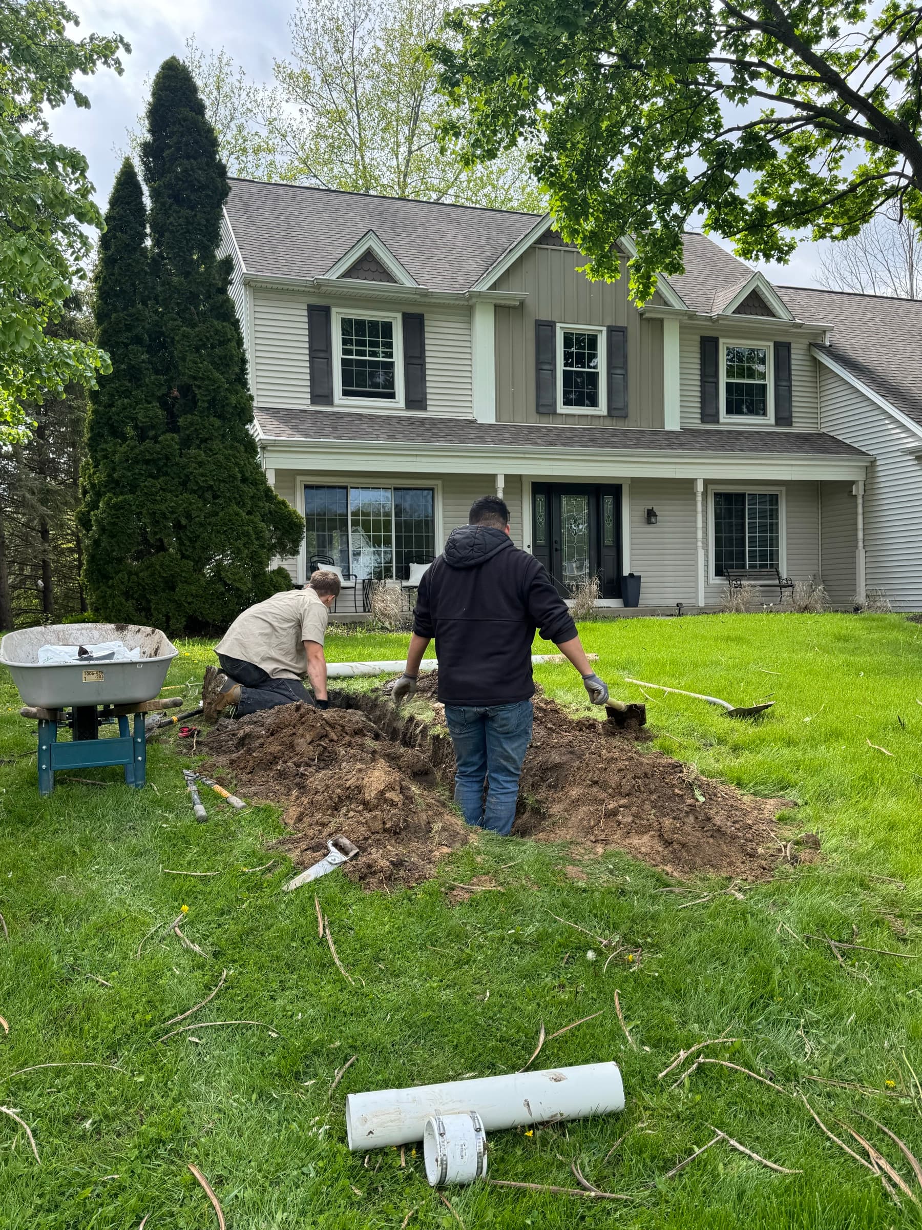 Crew trenching across the front lawn of a two-storey colonial with pipe staged ready