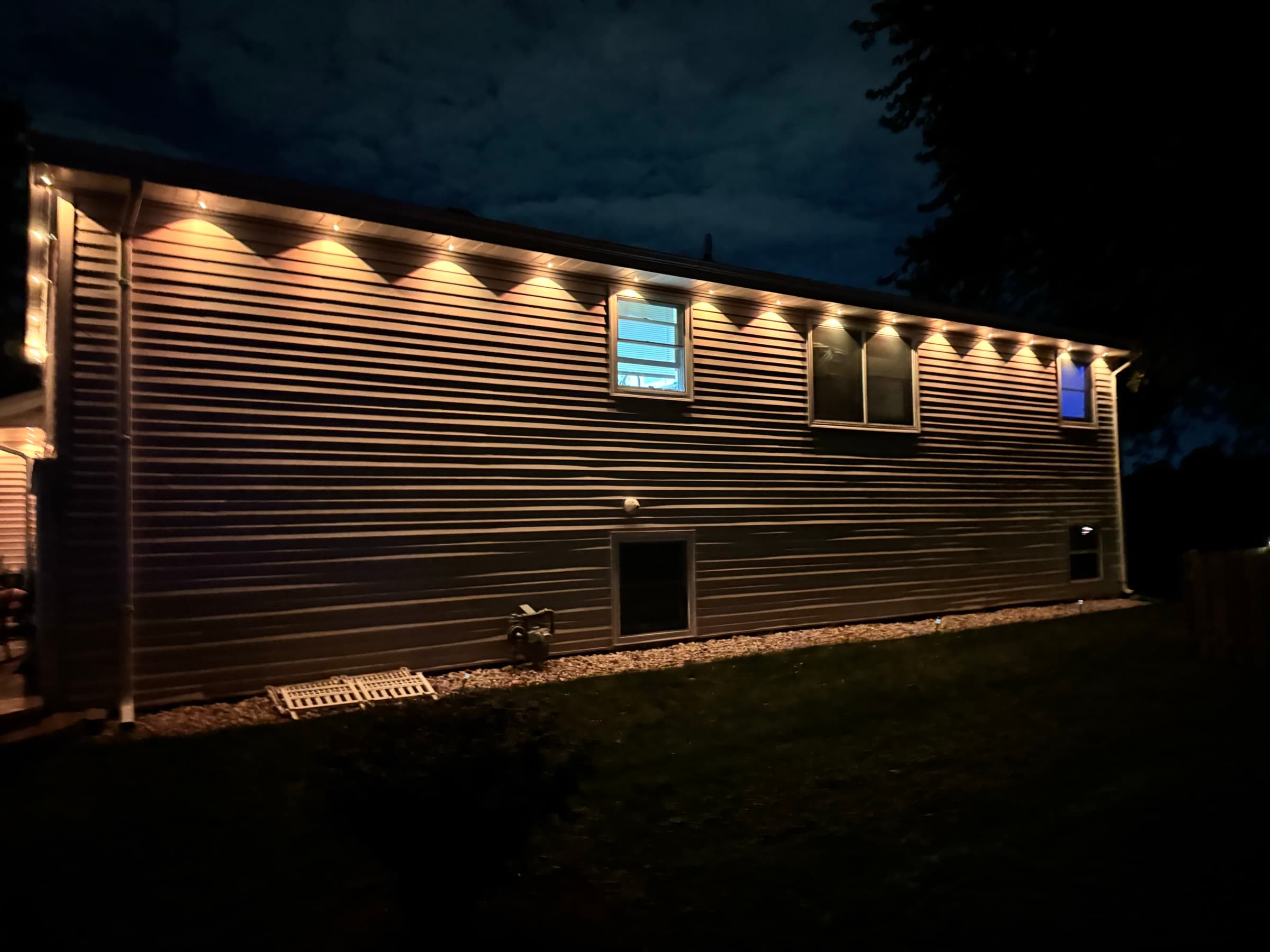 A long run of permanent lighting along the side of a house, each fixture throwing an even scallop of warm light down the wall