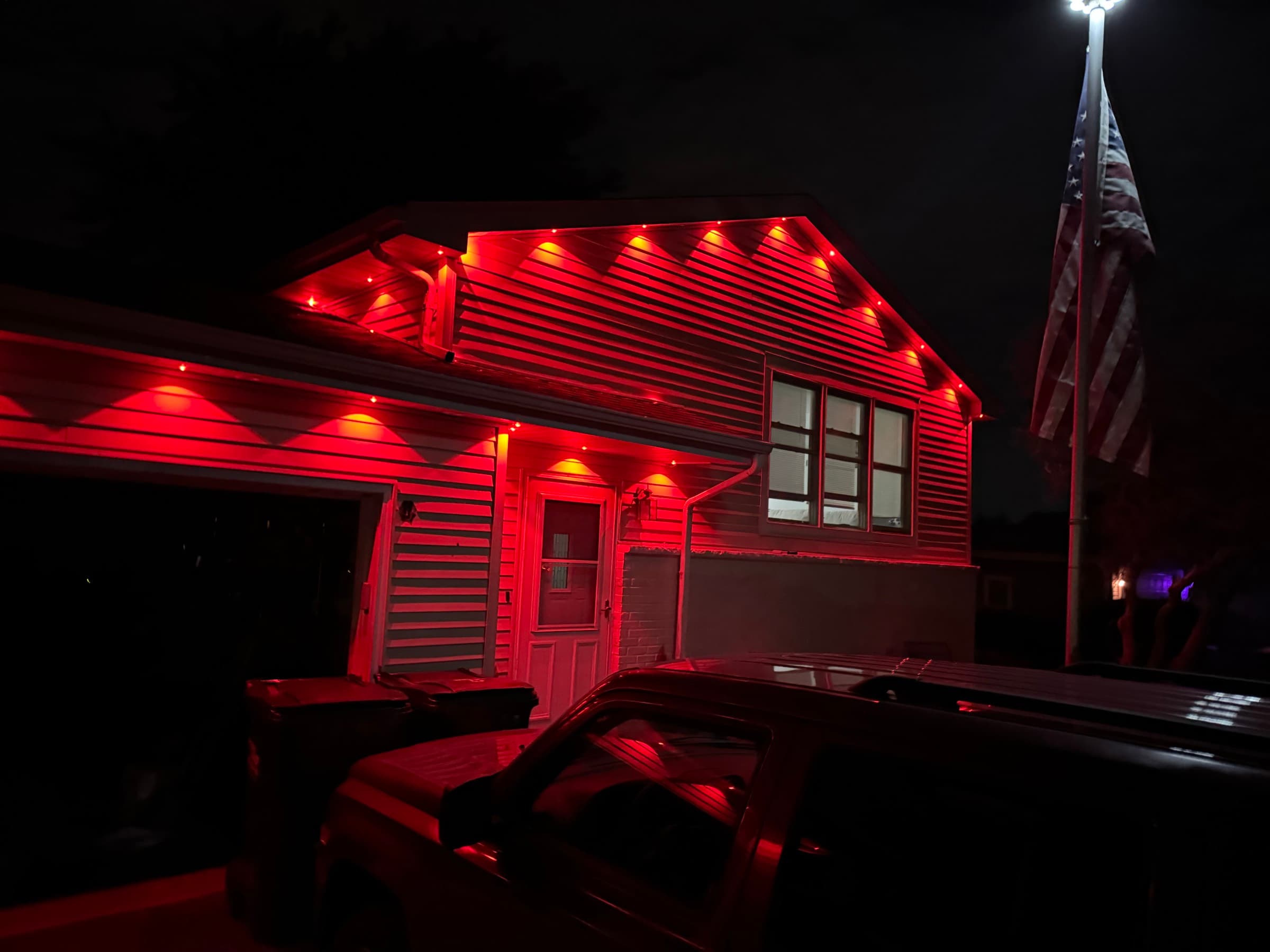 Red permanent lighting picking out the gable and garage line of a house at night, flag lit alongside
